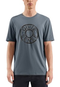 Mężczyzna w niebiesko-szarym t-shircie z krótkim rękawem z przetartym okrągłym logo z napisem "CISZA ZMYSŁÓW" z przodu, w zestawieniu z ciemnymi spodniami.