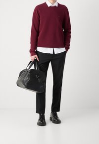 Pull bordeaux sur une chemise blanche, pantalon noir, chaussures en cuir noires, et un sac de voyage noir à double poignée. Textures lisses, design décontracté.