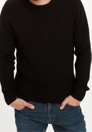 Mand iført en sort strikket sweater og blå jeans med hænder delvist i lommerne, beskåret ved skuldre og midt på lårene.