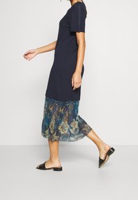 Granatowa sukienka z krótkimi rękawami, z przezroczystą nakładką w kwiatowy wzór. Zawiera teksturowane akcenty na rękawach oraz płaskie sandały.