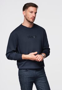 Homme portant un sweat-shirt bleu marine avec le texte "EXIMIO", un jean foncé et une montre argentée, debout les mains jointes devant un fond gris.