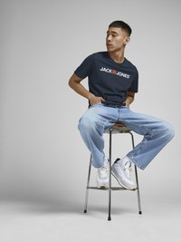 Jūrinis marškinėliai su "JACK & JONES" logotipu, šviesiai mėlyni laisvi džinsai ir balti sportbačiai. Modelis sėdi ant metalinio suolo prieš paprastą fono.