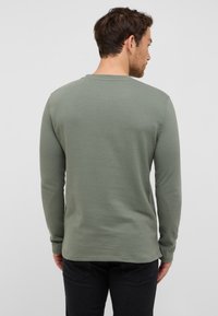 Koszula z długim rękawem w przytłumionym zielonym kolorze, teksturowana tkanina, okrągły dekolt i prosta dolna krawędź. Widok z tyłu pokazuje prosty wzór i brak wyraźnych akcentów.