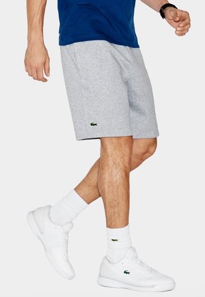 Shorts in cotone grigio con vita elastica, con logo Lacoste verde. Abbinati a scarpe da ginnastica bianche e calzini alla caviglia.