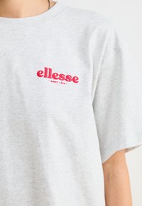 Šedé bavlněné tričko s kulatým výstřihem a krátkým rukávem, s červeně vyšitým logem "ellesse" a textem "since 1959" pod ním.