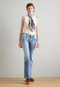 Jeans en denim léger, coupe skinny, bleu clair, légèrement évasés aux chevilles, associés à un haut sans manches crème et à une écharpe multicolore à motifs.