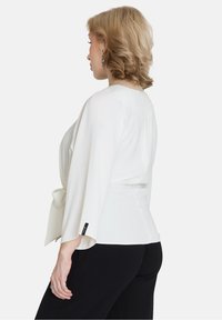 Blusa bianca con maniche ampie, vita legata e scollo rotondo. Tessuto liscio con una leggera lucentezza. Abbinata a pantaloni neri.