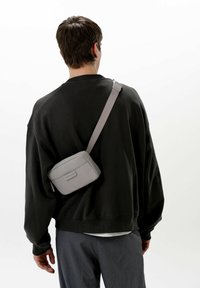 Šedá krosová taška obdélníkového tvaru a hladké textury, zdobená jemnou značkovou visačkou a nastavitelným popruhem. Nositelná přes tmavý sweatshirt.