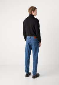 Juodas golfas, mėlyni laisvo kirpimo džinsai su užpakalinėmis kišenėmis, odinis užklotas ant juosmens diržo ir juodi batai. Paprastas fonas.