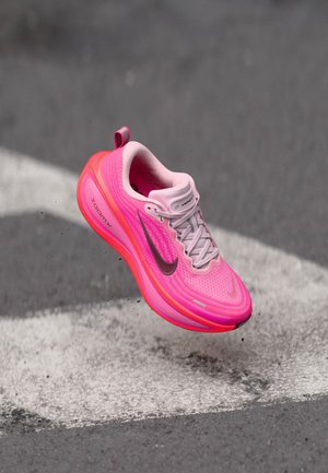 Zapatilla de correr Nike color rosa brillante con el logo Swoosh gris y suela ZoomX, suspendida sobre un asfalto gris con una marca vial blanca.