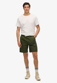 Pantalones cortos verdes con cinturilla elástica y cordón, combinados con una camiseta blanca. Se llevan zapatillas altas beige y calcetines a rayas.