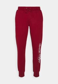 Vinsticka joggerbyxor i vinröd med dragsko i midjan och vit "Tommy Hilfiger EST.1985" logotyp tryckt vertikalt på vänster ben.