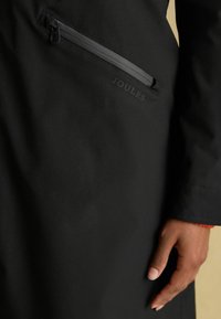 Jachetă neagră cu un buzunar cu fermoar, având un aspect neted și logo-ul "JOULES". Materialul este elegant, cu o aparență ușor texturată.