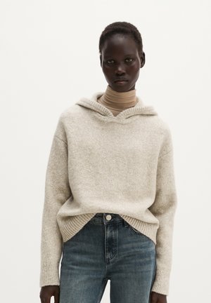 Junge Person mit kurzen Haaren, die einen beigen Rollkragenpullover unter einem hellgrauen gestrickten Hoodie und blaue Jeans vor einem schlichten weißen Hintergrund trägt.