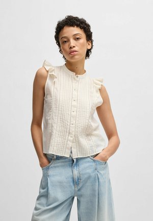 Blusa bianca senza maniche con spalle arricciate, pieghe verticali e chiusura con bottoni, abbinata a jeans a gamba larga blu chiaro.