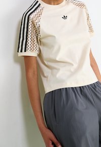 Krämfärgad t-shirt med korta ärmar, svarta randiga detaljer och spetsliknande utsmyckningar på ärmarna, med en liten svart logotyp på bröstet.