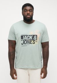 Light green cotton t-shirt with short sleeves, features graphic text "JACK & JONES" and tropical design accents in black and yellow.