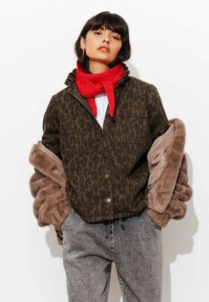 Veste imprimée léopard en tissu brun et vert foncé, doublée de fausse fourrure marron clair. Le modèle porte une écharpe rouge vif et un pantalon gris.