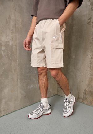 Beige shorts with elastic waistband and side pockets, featuring a small white logo. Paired with white sneakers with gray and red accents.
