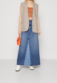 Beige ärmelloser Blazer über einem orangefarbenen gemusterten Crop-Top, kombiniert mit einem blauen Jeans-Maxikleid mit Schlitz vorne und weißen Knöchelstiefeln.