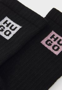 Calze di maglia nere con due loghi: toppe rettangolari bianche e rosa con "HUGO" ricamato in lettere in grassetto. Design testurizzato e a coste.