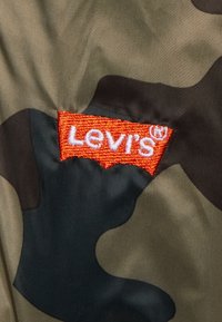 Logo ricamato Levi's in arancione su un tessuto camouflage con sfumature di verde e marrone, caratterizzato da una texture liscia e sintetica.