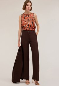 Top senza maniche arancione e nero a spirale abbinato a pantaloni scuri marrone high-waisted. Modella tiene un giubbotto abbinato. Tessuto morbido, gambe larghe.