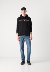Felpa nera con scritte bianche, jeans blu e sneakers nere. La felpa presenta un cordino e dettagli con logo sul davanti.