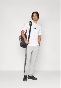 Ανδρικό μοντέλο με λευκό T-shirt με μαύρο λογότυπο, γκρίζες φόρμες με πλαϊνές ρίγες και μαύρα παπούτσια. Κρατάει μια μαύρη τσάντα Adidas.