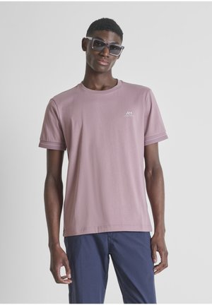 Mladić u svijetloplavoj majici kratkih rukava i tamnoplavim hlačama, s velikim kvadratnim sunčanim naočalama, stoji ispred jednostavne svijetle pozadine.