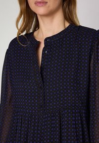 Svart blus med ett blommönster i blått och lila. Har en rund halsringning, knäppning fram och sammanknuten midja. Genomskinligt tyg.