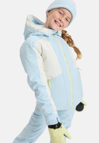 Lichtblauwe en crème ski-jack met gele accenten, voorzien van een ritssluiting aan de voorkant en zakken. Gecombineerd met gele handschoenen en een lichtblauwe muts.