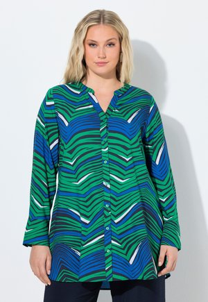 Kvinde iført en langærmet, knappet skjorte med et abstrakt stribet mønster i blå, grøn, sort og hvid, stående foran en ensfarvet baggrund.