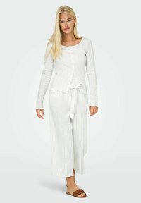 Ensemble de loungewear en tricot côtelé blanc comprenant un cardigan à boutons et un pantalon à cordon avec de subtils détails rayés. Des sandales plates complètent le look.