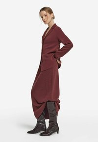 Burgunderfarbener Strickcardigan mit Kragen, doppelreihigen Knöpfen und strukturiertem Stoff. Kombiniert mit enganliegenden braunen Stiefeln bis zum Knie.