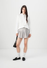 Chemise blanche à manches longues, jupe plissée grise, chaussettes blanches montantes aux genoux et mocassins noirs. Petit sac à main noir met en valeur la tenue.