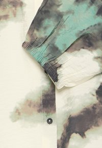 Manche de chemise boutonnée avec poignet élastique, présentant un motif camouflage style aquarelle en vert, beige et noir sur un tissu léger.