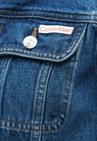 Keskmise sinise tooniga teksajakk, millel on sile pind. Disainis on Calvin Kleini silt, hõbedane pressnupp ja õmbluselemendid.