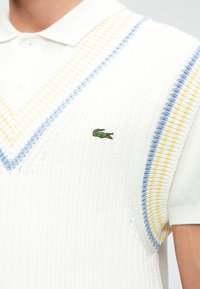 Λευκό πλεκτό αμάνικο πόλο πουλόβερ με μπλε και κίτρινες ρίγες. Διαθέτει V-λαιμόκοψη και μικρό πράσινο λογότυπο κροκοδείλου στο στήθος.