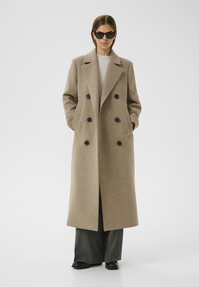 Abrigo de lana marrón claro de doble botonadura con grandes botones negros, bolsillos laterales y solapas de muesca, combinado con pantalones amplios grises y zapatos negros.