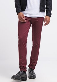 Burgundské slim-fit kalhoty vyrobené z pružné tkaniny, s hladkou texturou a minimem detailů, doplněné černými šněrovacími botami.