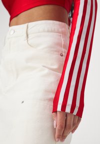 Κόκκινο μακρυμάνικο μπλουζάκι με τρεις λευκές ρίγες στα μανίκια, συνδυασμένο με λευκά τζιν παντελόνια, τα οποία διαθέτουν μπροστινή τσέπη και λεπτομέρεια από κουμπί.