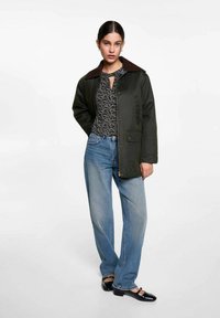 Donkergroene jas met een bruine corduroy kraag, knoopsluiting en zijzakken. Draagt over een gestreept topje en gecombineerd met lichtgewassen jeans.