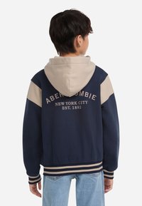 Felpa con cappuccio blu navy con accenti beige, polsini e orlo a coste, con la scritta "Abercrombie New York City EST. 1892" in lettere color crema.