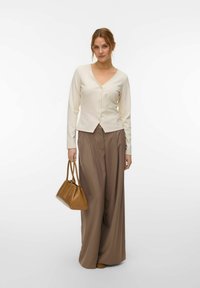 Femme portant un haut crème à boutons et un pantalon large marron, tenant un sac à main beige, debout devant un fond blanc uni.