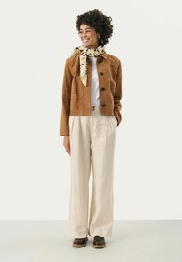 Bruine suède jas met zwarte knopen, witte t-shirt en lichtbeige wijde broek. Patroonsjaal om de nek geknoopt. Bruine schoenen.