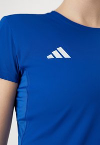 Blå kortärmad träningsskjorta med vitt Adidas-logotyp på bröstet, bärs av en person som visas från halsen till överarmen.