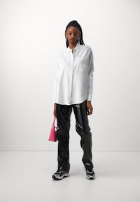 White long-sleeve shirt with a single pocket, glossy black pants, and a small pink handbag. Sleek sneakers complete the look.