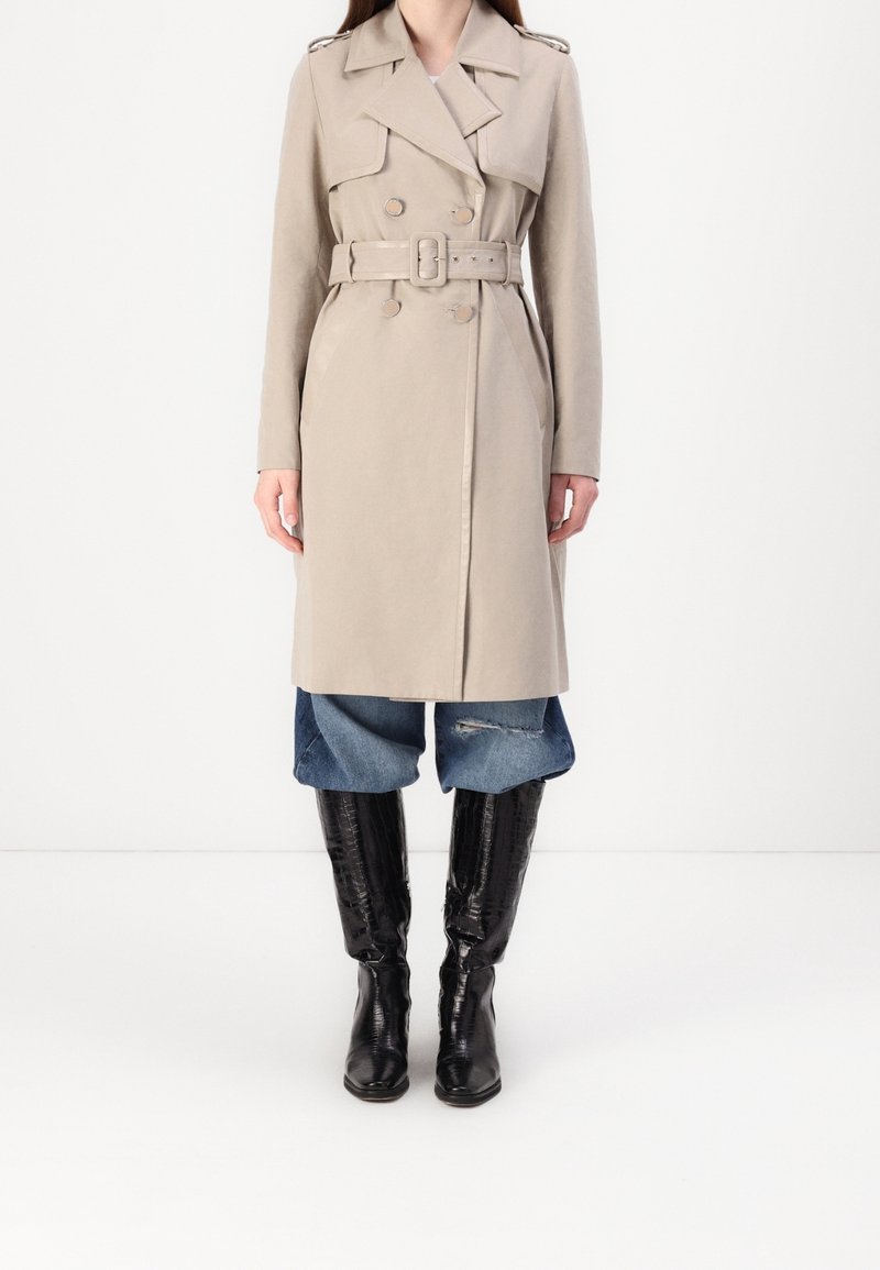 Femme portant un trench-coat beige ceinturé sur un jean bleu retroussé et des bottes noires montantes, debout devant un fond blanc uni.
