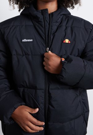 Persona con pelle scura che chiude la zip di una giacca a piumino nera Ellesse con loghi del marchio sul petto e sul polsino della manica.